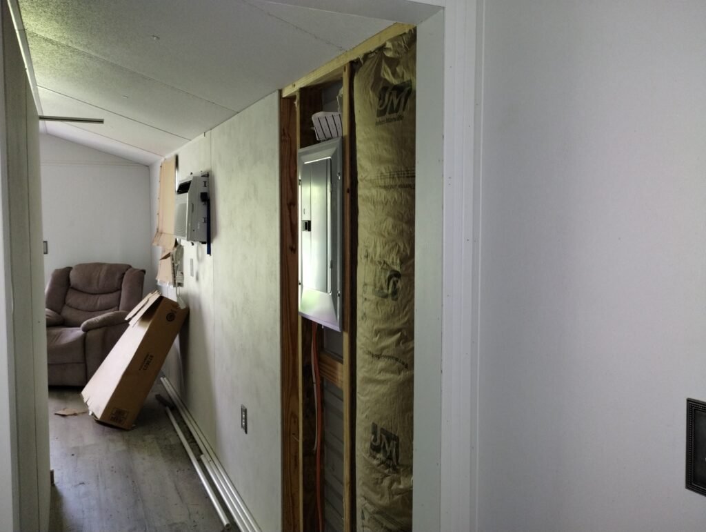 view looking back down the hallway towards the sitting room the electric panel is installed in the wall, insulation is exposed, air conditioner is in the wall, a brown recliner is in the sitting room corner with a cardboard box on the floor in front of it