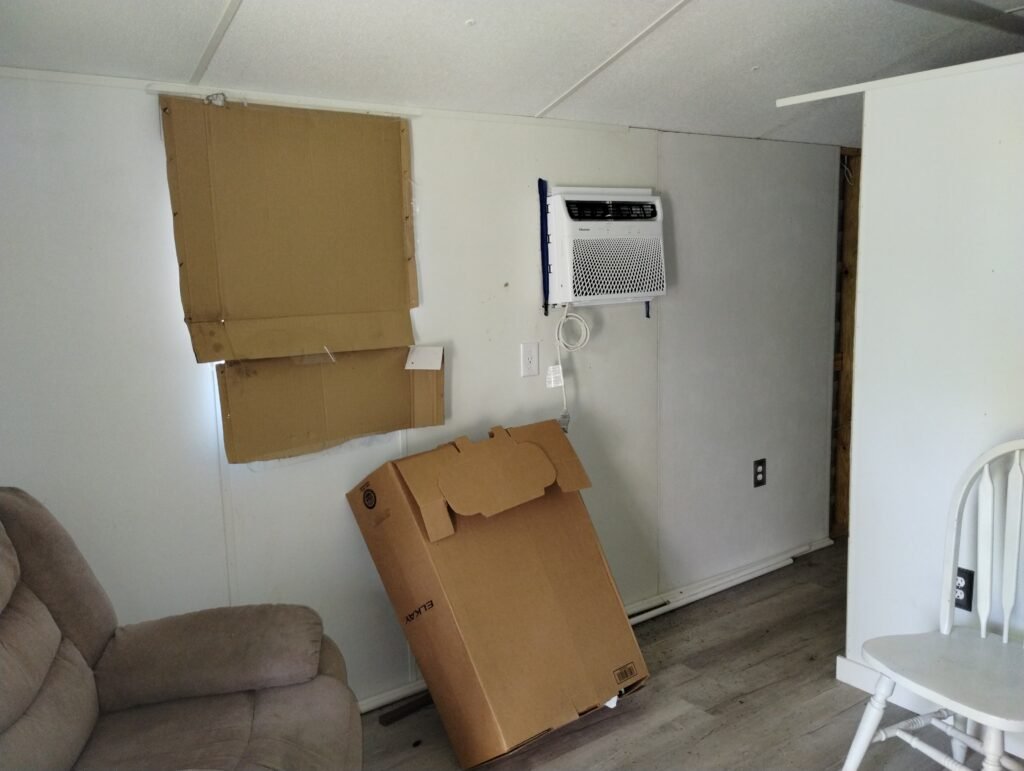 view from the front door looking towards the left, part of a recliner is showing in the bottom left of the image, a cardboard box is leaning against the wall, the window is covered with cardboard and there is a new air conditioner in the wall just past the window