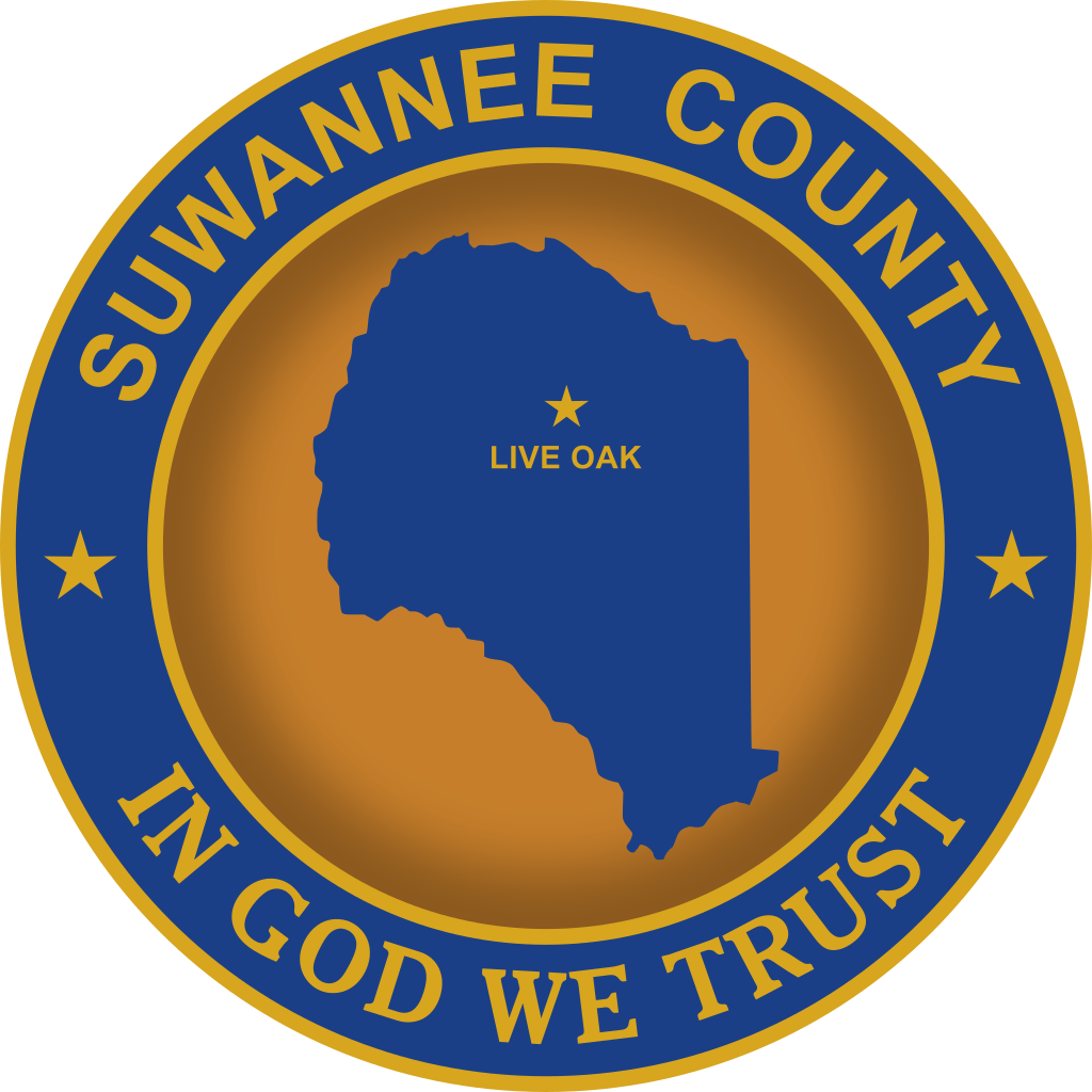 Suwannee County Seal with the shape of Suwannee county in dark blue on a golden background . The city of Live Oak is marked in gold .  The words Suwannee County and In God We Trust are gold, printed on a ring of blue surrounding the center golden circle with the blue shape of the county.